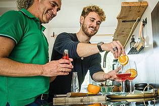 Christin Klose/dpa-tmn | Freude am Experimentieren: Freunde bereiten in der heimischen Küche einen alkoholfreien Aperitif mit Sanbitter zu. Er ist für seine rote Farbe und bittersüße, herbe Geschmacksnote bekannt.