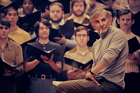 Filmszene: Leonard Bernstein in Maestro sitzt vor einem erwartungsvollen Chor. 